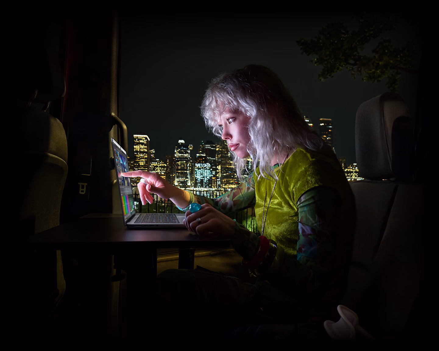 A person sitting in the back seat of a van, the open passenger door reveals a city skyline at night, MacBook Pro with PROLED XDR Display on a worktable, the person is illuminated by the screen while using the Touch feature to edit a video in DaVinci Resolve Studio